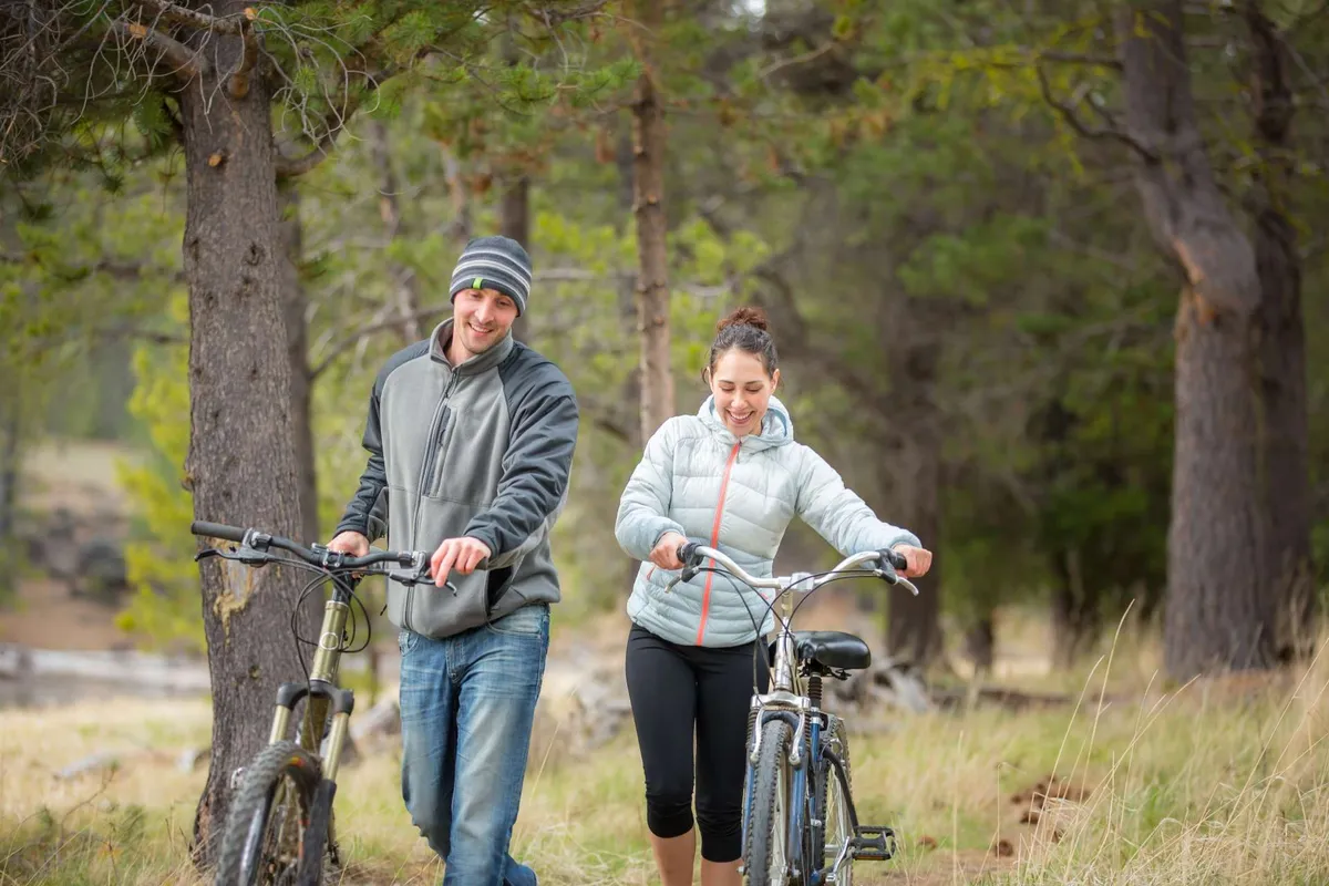 Two cyclists walk their bikes through a sunlit, grassy area bordered by tall trees, suggesting easy access to outdoor trails and a blend of urban greenery. The relaxed, joyful mood reflects the spirit of adventure and the accessibility of nature in Bend.