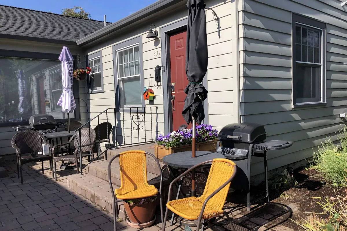 A cheerful, sunlit patio featuring a bold red door as the backdrop, inviting yellow Adirondack chairs, a gleaming gas grill, and vibrant flower pots. The space is set up for both outdoor dining and relaxing, with all the essentials for social gatherings or peaceful solo moments.