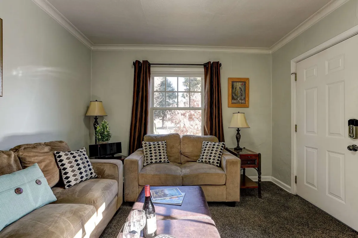 A comfortable living room scene with a sofa adorned with decorative pillows, a table with wine and glasses, natural light streaming through the window, and tasteful artwork on the wall. The ambiance suggests intimate gatherings or quiet evenings spent in comfort.