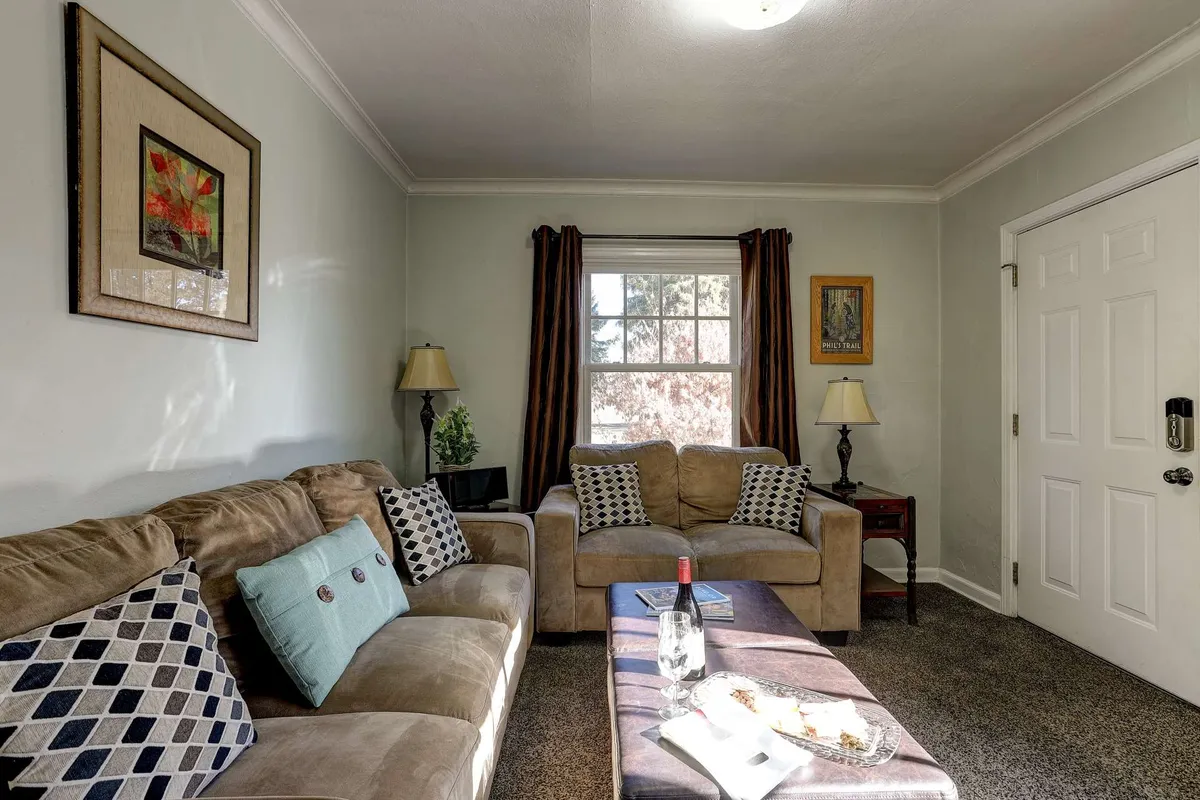 A cozy living room featuring a plush sofa set beside a coffee table, with artwork and lamps adding warm accents. Large windows let in natural light, enhancing the intimate ambiance. The scene suggests a space perfect for relaxing, reading, or enjoying a quiet evening, with the television visible for entertainment.