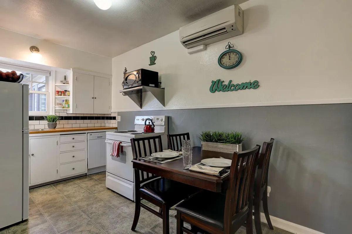 A bright, inviting kitchen scene featuring a cozy eat-in table with chairs, a stove and oven, an open fridge, a decorative clock, and a welcoming sign. Sunlight fills the room, creating a cheerful and homey ambiance ideal for cooking and sharing meals.
