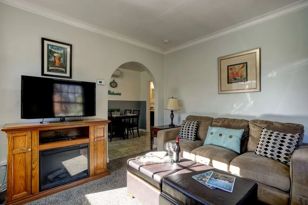 The image showcases a warm and inviting living room space inside Drake Cottage 311. It features a plush sofa facing a modern television, a stylish coffee table, ambient lighting from a nearby lamp, and thoughtfully chosen artwork on the walls. The overall atmosphere is cozy and intimate, with tasteful decor that invites relaxation and leisure.