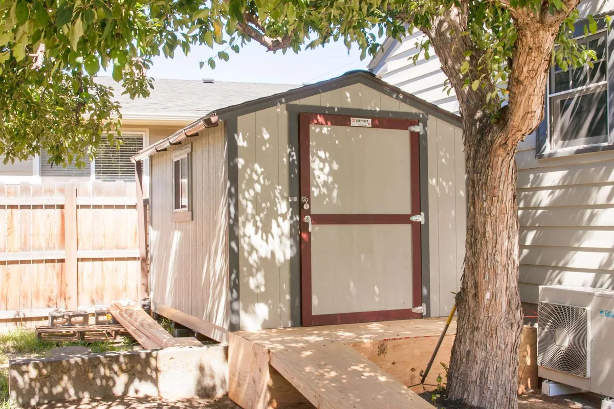 A functional exterior scene highlighting a cozy storage shed nestled beneath a leafy tree, with a wooden ramp leading up to it. The setup hints at practical storage for outdoor gear while maintaining visual harmony with the surrounding outdoor space.