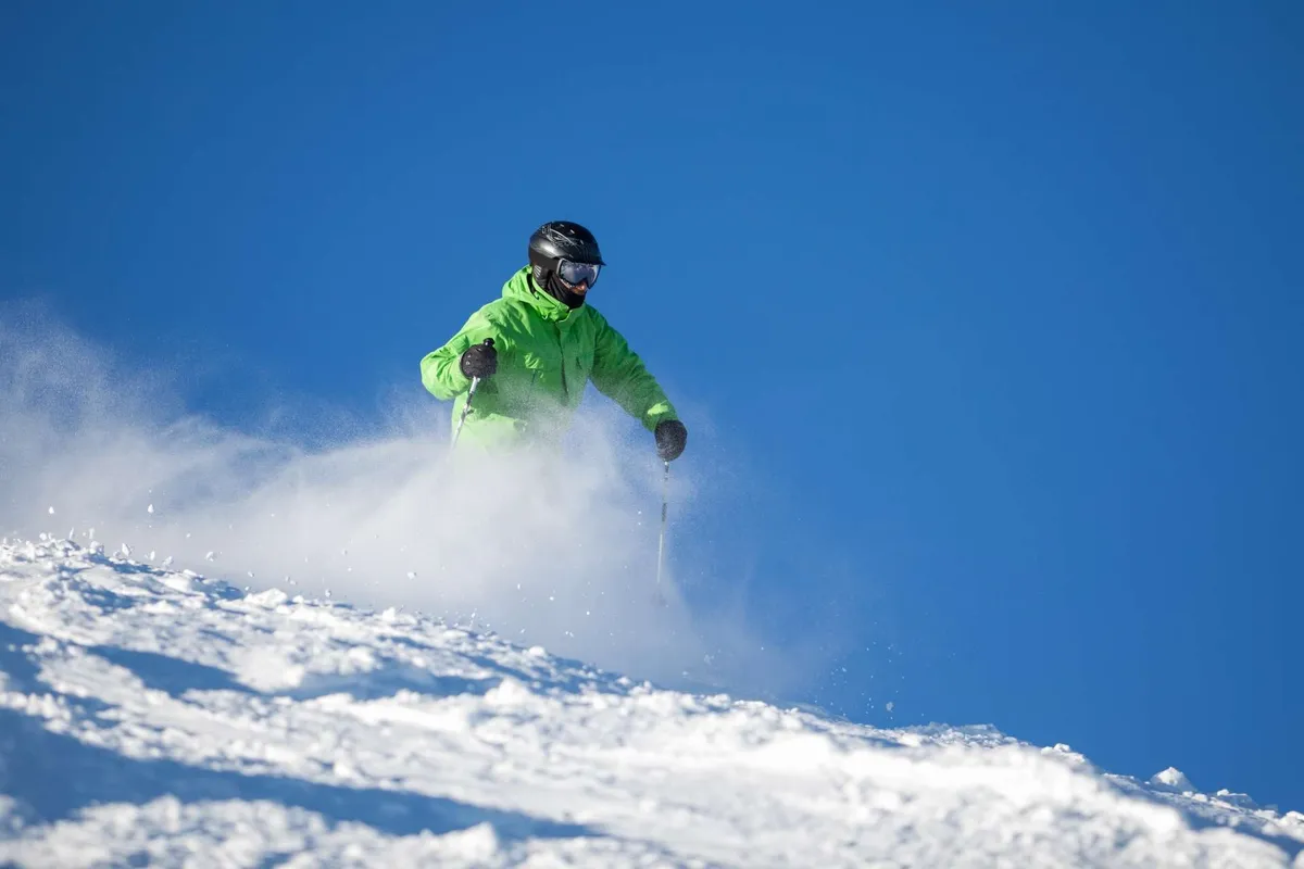 A skier in a vibrant green jacket carves down a snowy slope under a clear blue sky, with untouched snow all around. The image radiates energy and adventure, perfectly capturing the thrill of outdoor sports in winter.