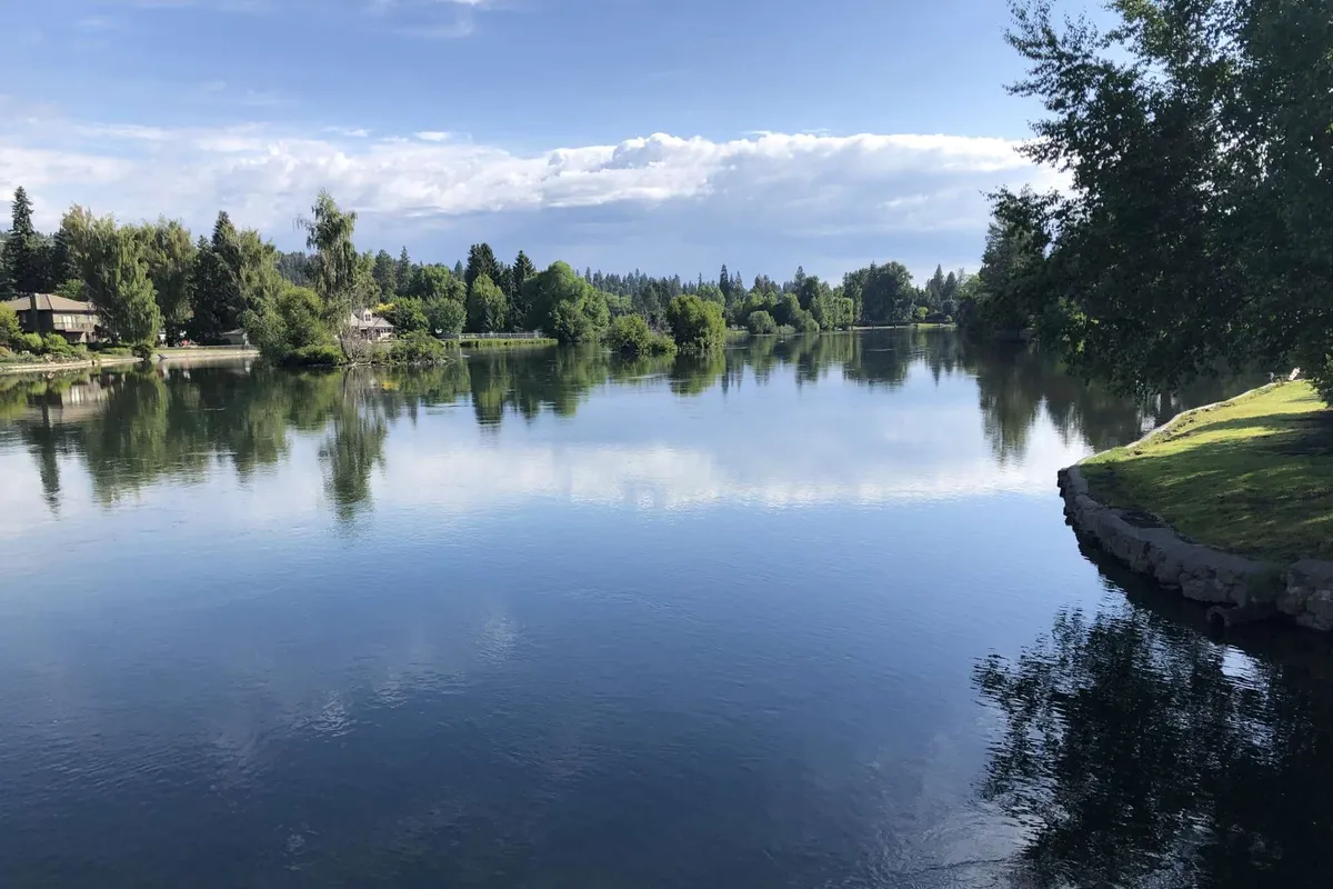 A tranquil scene of calm water with lush greenery and distant hills, evoking the peaceful ambiance of Drake Park, which is mentioned as just across the street. The view hints at outdoor recreation, romantic walks, and the natural beauty that forms part of the downtown experience.