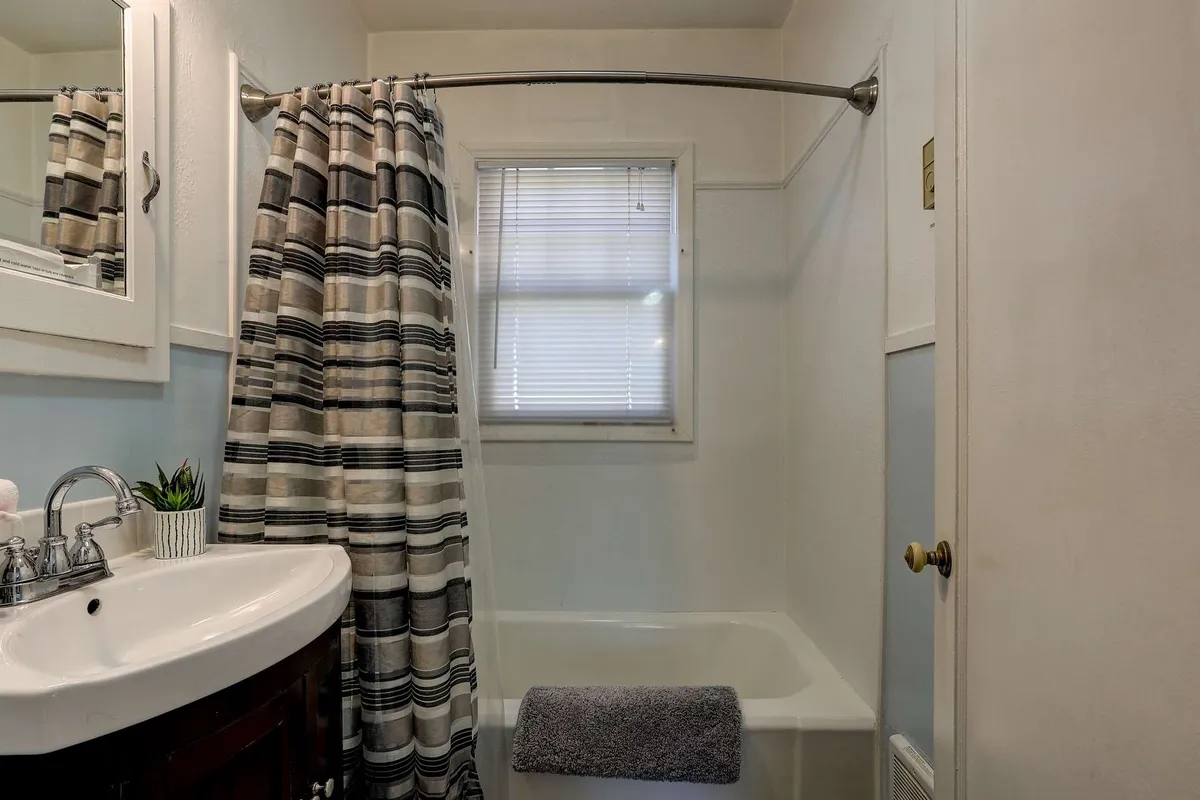 An updated bathroom with a clean sink, modern mirror, shower curtain, and window with blinds. The space feels serene and inviting, providing both a tub and shower for relaxing soaks or refreshing starts to the day.