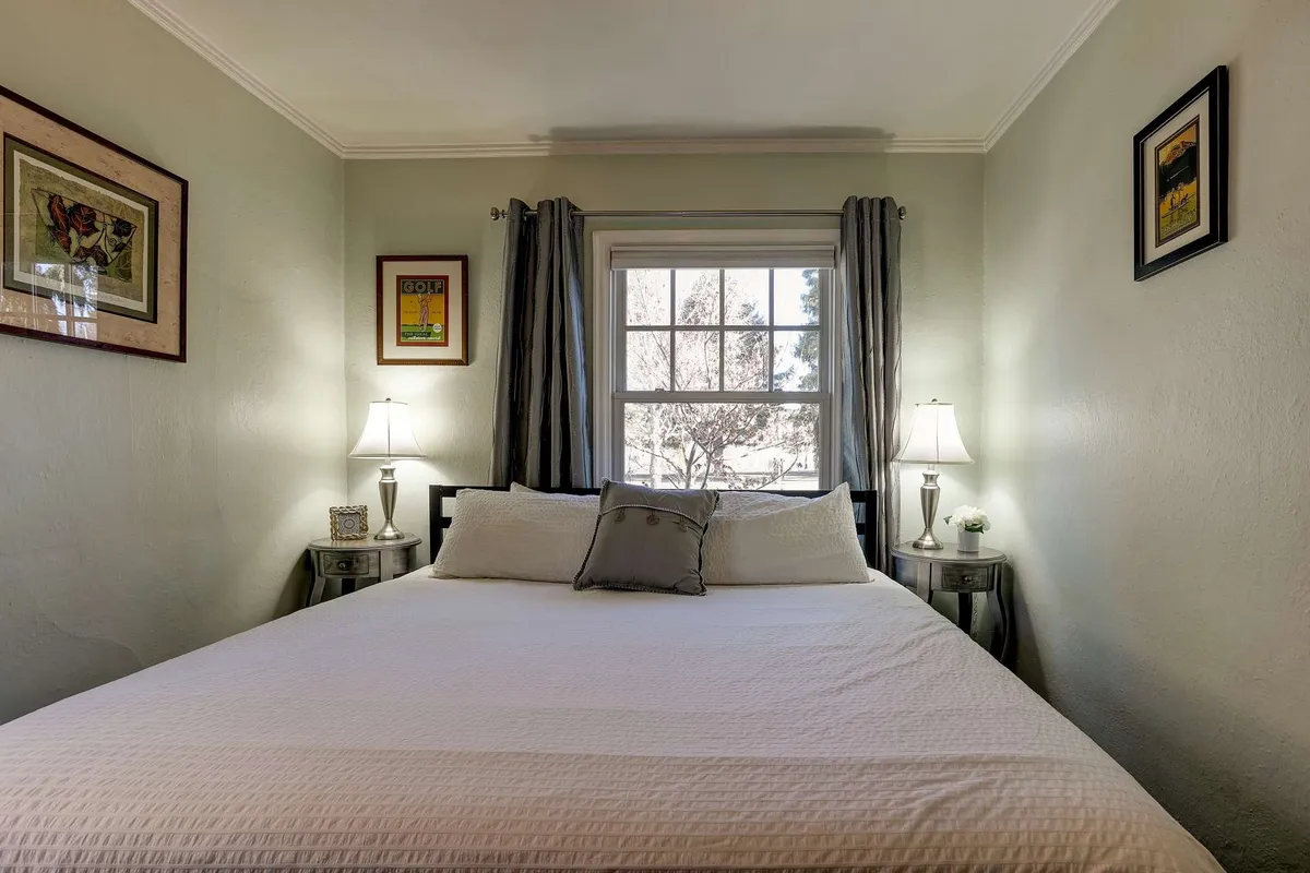 A spacious bedroom with a comfortable bed, glowing bedside lamps, a window letting in soft natural light, and artistic touches on the walls. The calm atmosphere invites reading or resting in peace.